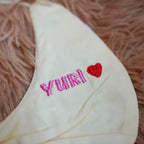 White baby bib with 'Yuri!' embroidered in pink on a fluffy brown surface